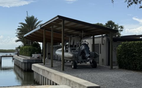 Boat stored on a trailer beneath a wooden shelter at a waterfront dock, with calm water on the left and palm trees in the background.