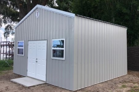 Gray metal storage shed with white double doors and two small windows, set on dirt with a concrete ramp in front and trees in the background.