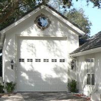 White two‑car garage with a circular window above the door, lantern-style lights, and a paved driveway framed by trees on a sunny day.