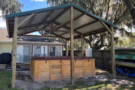 Wood-framed outdoor pavilion with a wooden hot tub under a metal roof, in a backyard near a house and boat storage area.