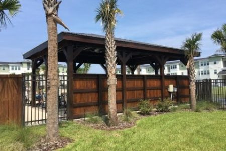 Wooden gazebo with a dark roof in a grassy yard, framed by palm trees and a brown fence.