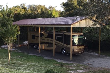 A large motorhome parked under a wooden carport with a metal roof in a grassy, tree-filled yard.
