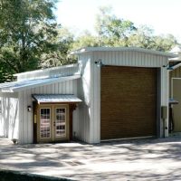 White metal storage building with a large brown roll-up door, a small glass-paned entry, and a second side door; paved driveway and trees in the background.
