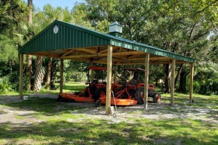 Open-sided wooden shelter with a green metal roof housing several orange riding lawn mowers outdoors among trees and grass.