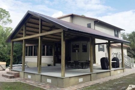 Two-story beige house with a large open wooden deck and covered patio supported by thick posts; outdoor dining table and grill on the deck.