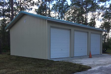 Metal three-bay garage with blue trim and white doors, set among pine trees; orange traffic cone on gravel driveway nearby.