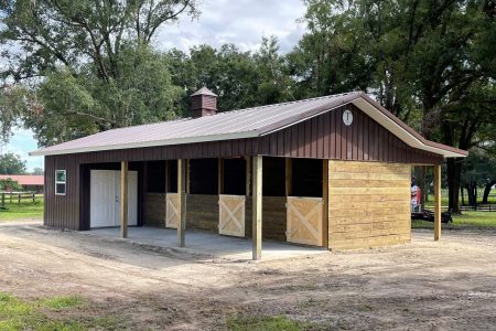 Front view of a brown wooden stable with open stalls under a metal roof and a small cupola, surrounded by trees in a rural setting.