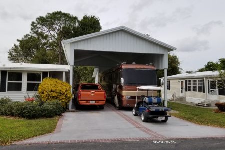 Carport attached to a white house with an orange SUV and a brown motorhome parked inside; a golf cart is in the driveway front.