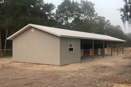 Gray metal shed with a white peaked roof and open-sided stalls, dirt ground in front, trees in background. -Side view of a gray metal building with a triangular roof and open shelter area supported by wooden posts. -Equestrian-style barn under an overcast sky, gray siding, dirt yard, and surrounding trees. -Small gray shed near a fenced corral with a row of trees behind. -Wood-framed open-sided barn with a metal roof and dirt yard.