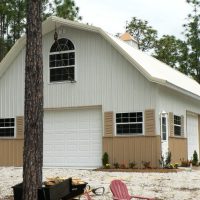 White barn-style garage with a tall arched window, beige lower siding, and pine trees surrounding.