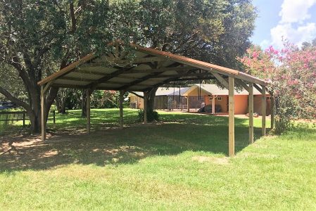 Wooden open-sided carport shelter in a grassy yard with trees and a house in the background