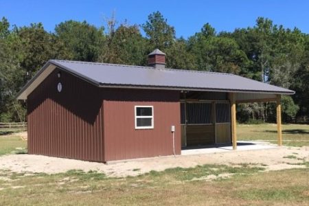 Brown metal-roofed shed with an open-sided shelter on the right, in a grassy, tree-filled area.