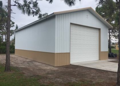 Blue metal storage shed with a white roll-up door and a tan bottom trim on a concrete pad, surrounded by trees in an open area.