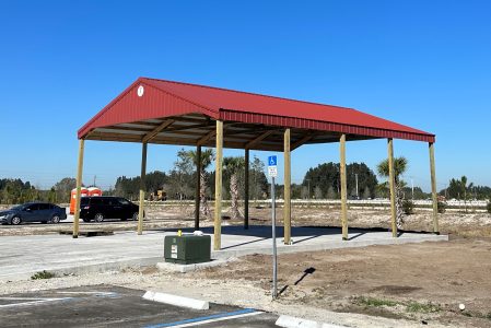 Open-air shelter with a red metal roof supported by wooden posts in a paved parking area under a clear blue sky; a blue handicap parking sign is visible nearby.