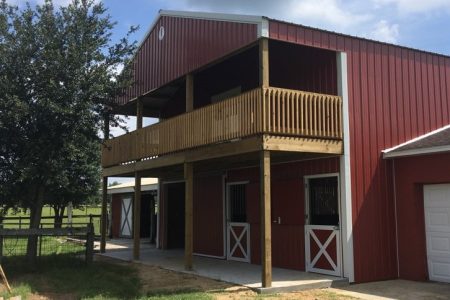 Red barn-style building with a wooden second-floor balcony, white X-panel barn doors on the ground level, set in a rural yard with a tree and fence.