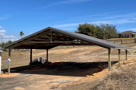 Open-sided wooden carport with a metal gable roof over a dirt lot; construction materials lie beneath and suburban houses are in the background under a blue sky.