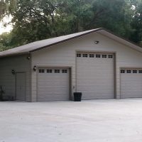 Detached three-car garage with a large central door and two smaller side doors on a concrete driveway, surrounded by trees.