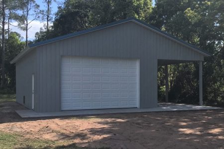 Gray metal garage with a large white sectional door and a right-side open carport, set among trees on a dirt foreground.
