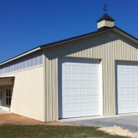 Beige metal outbuilding with two large white garage doors, a small cupola on the roof, and a concrete driveway.