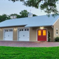 Beige four-bay garage with white doors and a red front door on a brick driveway, surrounded by green lawn and trees in the background.
