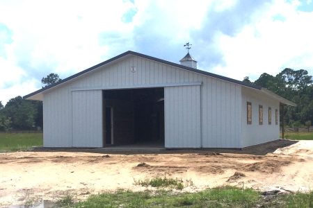 White metal barn with a large open doorway, small cupola and weather vane, in a rural grassy field under a blue sky.