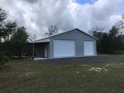 Gray metal storage building with two large white roll-up doors on a gravel lot under a cloudy sky.