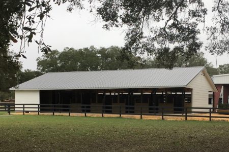 White, single-story barn with a metal gable roof and open stalls, bordered by a dark wooden fence in a grassy field.