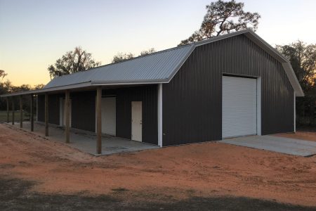 Long dark corrugated-metal building with a peaked roof and white trim, large roll-up door, set on a dirt lot at sunset