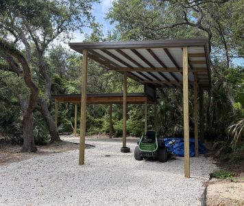 Open-sided wooden shelter with a corrugated metal roof in a wooded yard; gravel ground and a green riding mower parked underneath, with blue tarp nearby.