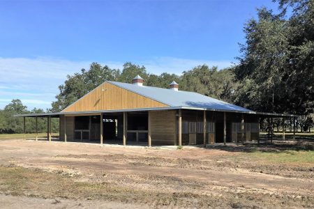 Wooden barn with a silver metal roof and two cupolas, open stalls at the front amid trees outside.