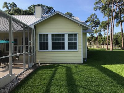 Yellow house with a large white-framed window; screened porch on the left and a green lawn in the foreground.