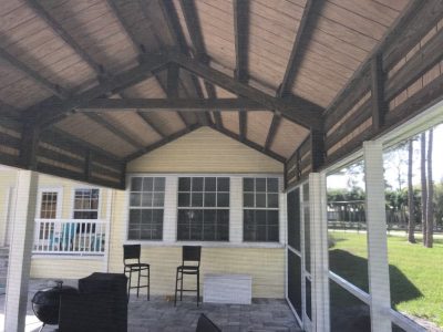 Screened porch with exposed wooden beams overlooking a yard; yellow house with large window and two bar-height chairs on a tiled patio.