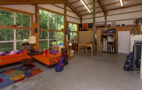 Spacious artist studio with exposed wooden beams, a bright orange sofa, colorful rug, and art supplies around a worktable by large windows.