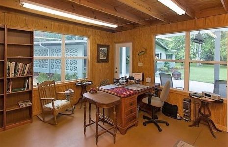 Wood-paneled home office with large wooden desk, rolling chair, and shelves; windows reveal a green yard outside.