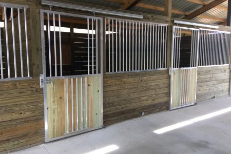 Row of wooden-and-metal horse stalls inside a barn, with vertical metal bars and latching doors along a wood-paneled wall.