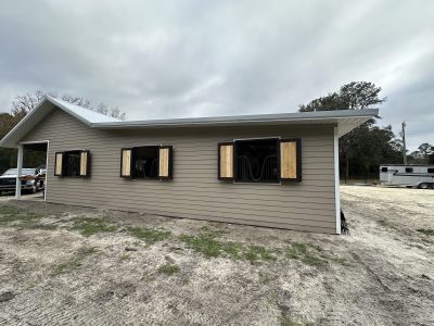 Two-tone beige modular building with open horse stalls and boarded wooden shutters under a cloudy sky, on dirt ground.