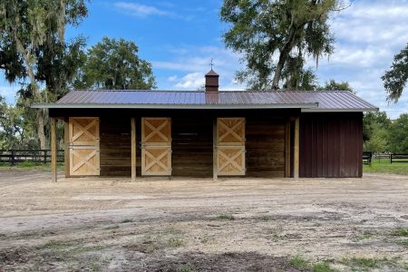 Rustic stable with four light wood doors under a blue metal roof, set in a dirt yard with trees in the background.