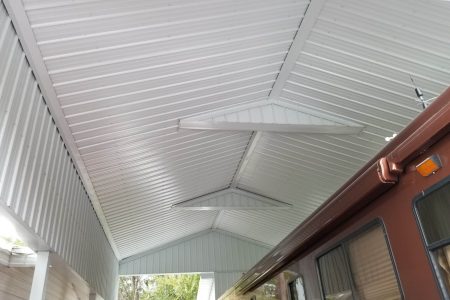 Interior view of a corrugated metal roof with white support beams forming triangular sections, overlooking a brown building wall with windows on the right and some greenery outside.
