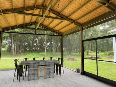 Covered patio with glass walls and a wooded backyard view; a stone bar island with metal stools beneath a wooden beam ceiling and hanging bulbs.