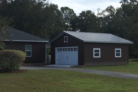 Detached brown garage with white trim and a white garage door, metal roof, in a grassy yard with trees behind.