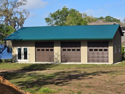 Garage-style building with a teal sloped metal roof and three brown panel doors against a blue sky and trees in the background.