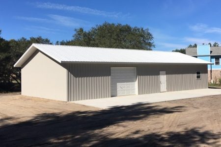 Long beige metal storage building with a white corrugated roof, large garage door, and a side door on a dirt lot under a blue sky.