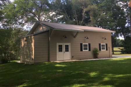 Beige outbuilding with a metal roof, white double doors, two small windows with black shutters, in a grassy yard.