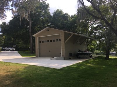 Residential driveway with a beige garage and carport, a boat trailer with jet skis parked under the shelter, surrounded by tall trees and green lawn.