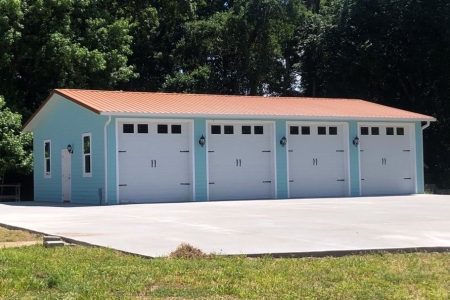Blue four-car garage with white doors and a red metal roof, set on a wide concrete driveway with trees in the background.
