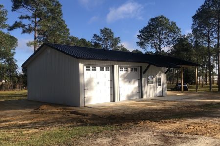 Grey metal garage with two white carriage-style doors, a side entry door with windows, and a black pitched roof set among tall pine trees; dirt driveway in front.