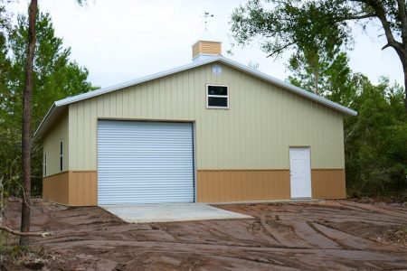 Beige metal storage shed with a large gray roll-up door and a small white entry door, surrounded by trees.