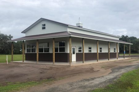 Long, white-and-brown, barn-style building with a wraparound porch supported by wooden posts under a cloudy sky.